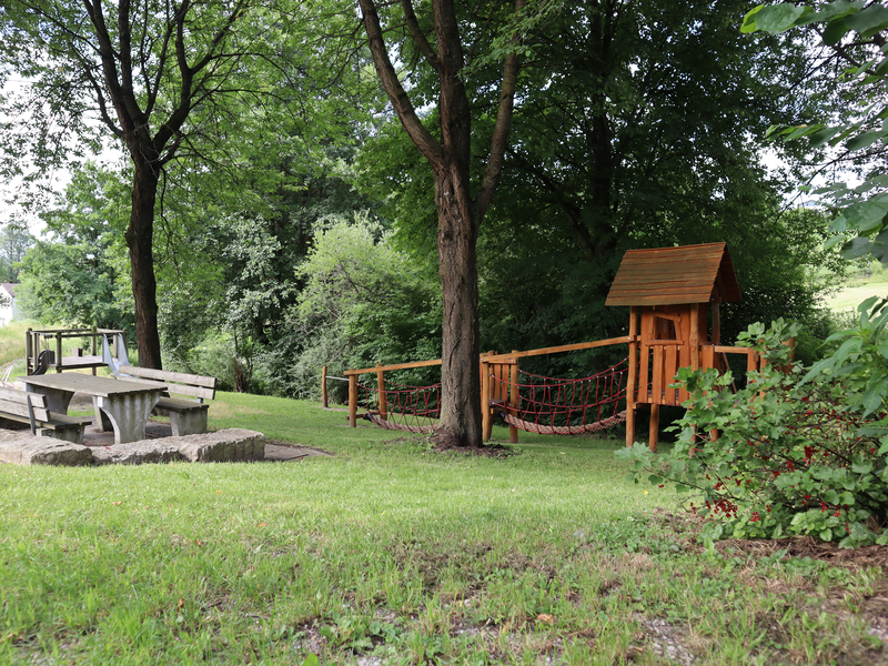 Ein Spielplatz mit Holzspielhaus mit Hängebrücke sowie ein Tisch mit Bännken umrundet von Bäumen. 