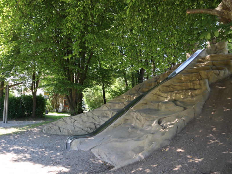 Eine lange Rutsche ist in einen künstlichen Felsen integriert, umgeben von schattenspendenden Bäumen in einem Park.