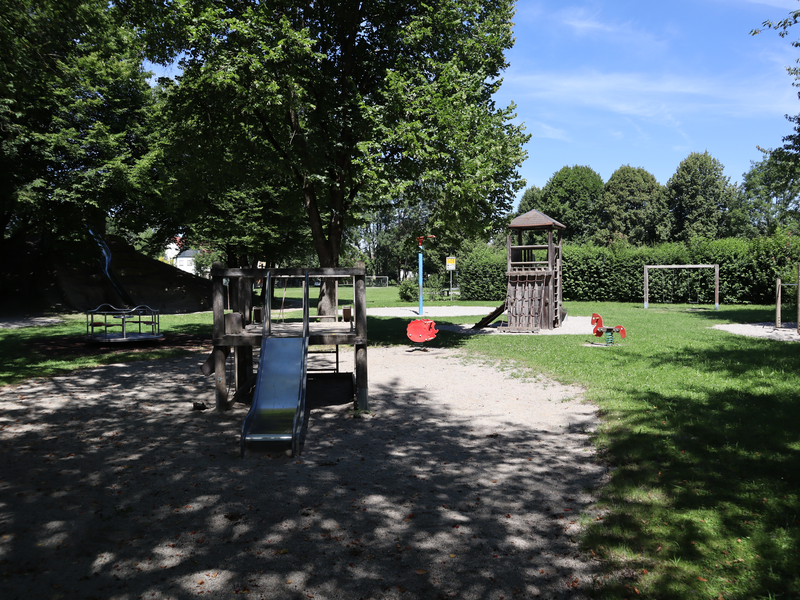 Ein sonniger Spielplatz mit vielen Spielgeräten, umgeben von Bäumen.