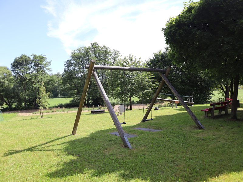 Ein weitläufiger Spielplatz mit Doppelschaukel im Vordergrund, umgeben von Bäumen.