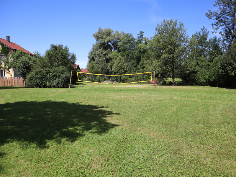 Auf einer gemähten Wiese umgeben von Bäumen und einem Haus, ist ein Volleyballnetz aufgestellt.