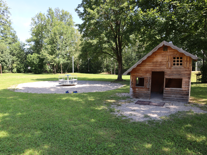 Ein Spielplatz mit einem Holzspielhaus und links daneben einem Karussell.