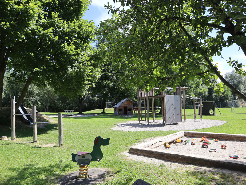 Ein Spielplatz mit vielen Spielgeräten wie Federwippe, Reck, Hangrutsche, Sandkasten und Kletteranlage umgeben von Bäumen