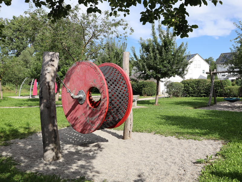 Ein Spielplatz im Freien mit einer großen, roten rotierenden Scheibe aus Metall ist von Gras und Bäumen umgeben, im Hintergrund sind einige Wohnhäuser zu sehen.