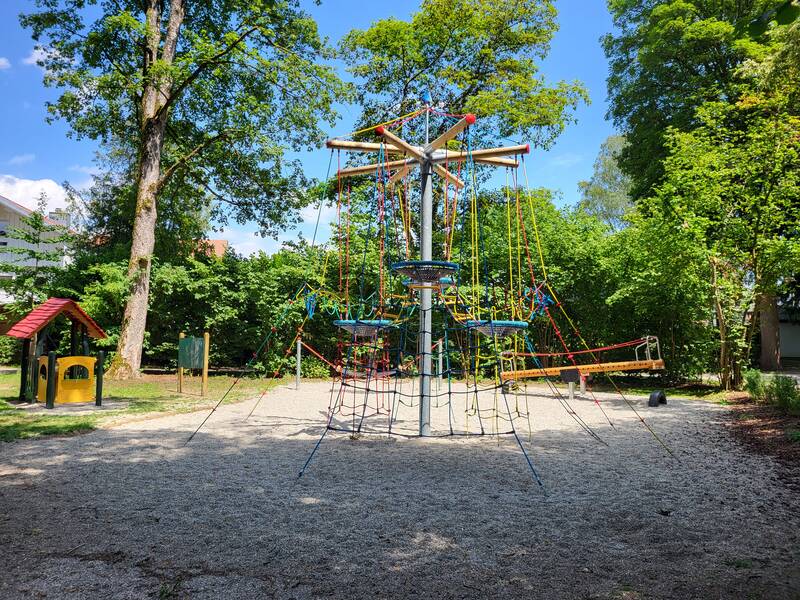 Ein Spielplatz miteinem Nestbaum im Mittelpunkt und vielen weiteren Spielgeräten in einer Parkanlage bie strahelnd blauem Himmel.