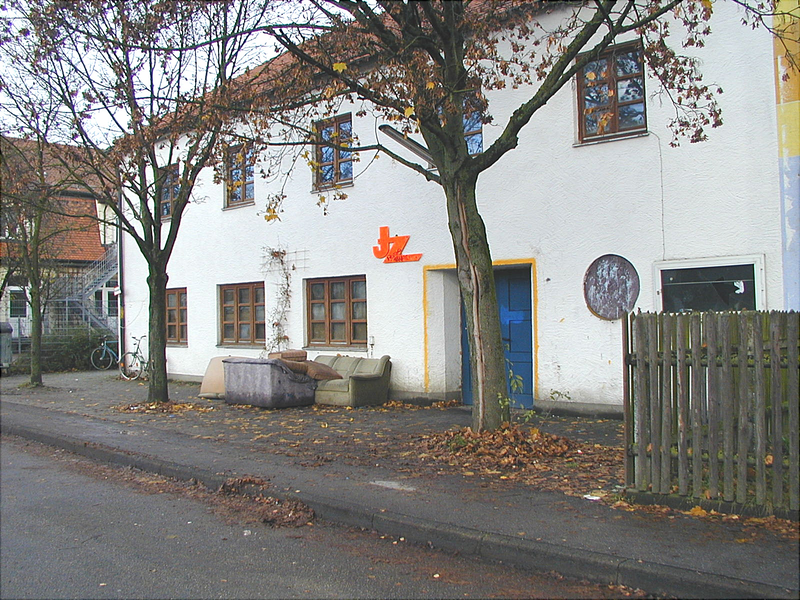 Ein weißes Gebäude mit mehreren Fenstern, einem blauen Eingangstor und einem Sofa, das auf dem Gehweg zwischen zwei Bäumen steht.