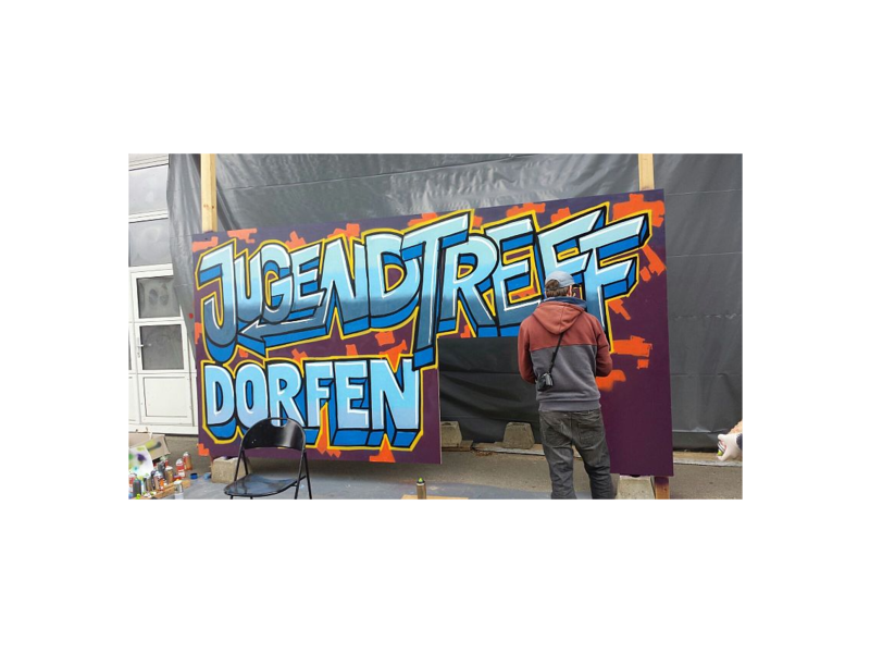 Ein Mann sprüht ein buntes Graffiti mit der Aufschrift "Jugendtreff Dorfen" auf eine Wand.