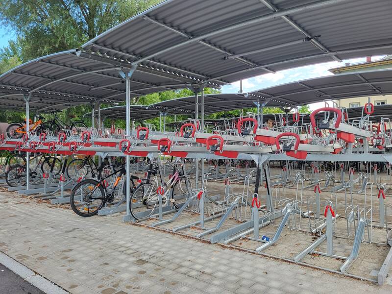 Ein überdachter Fahrradstellplatz mit zweistufigen Fahrradständern, von denen einige Fahrräder halten, ist zu sehen.