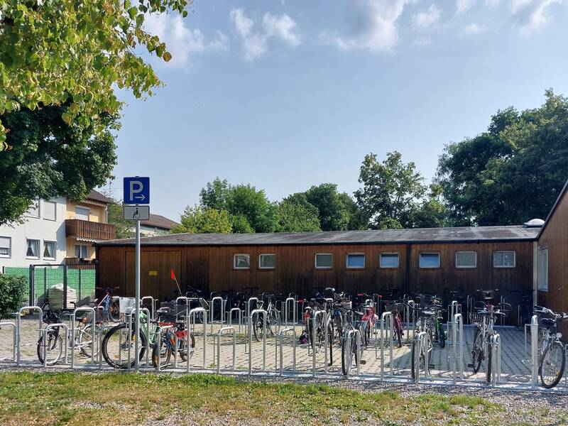 Vor einem schlichten Gebäude stehen zahlreiche Fahrräder in einem Fahrradständer, umgeben von Bäumen und einem Behindertenparkplatzschild.