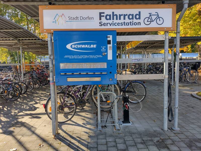 Eine überdachte Fahrradstation mit einem blauen Schwalbe-Automaten für Reparaturartikel steht neben vielen geparkten Fahrrädern.