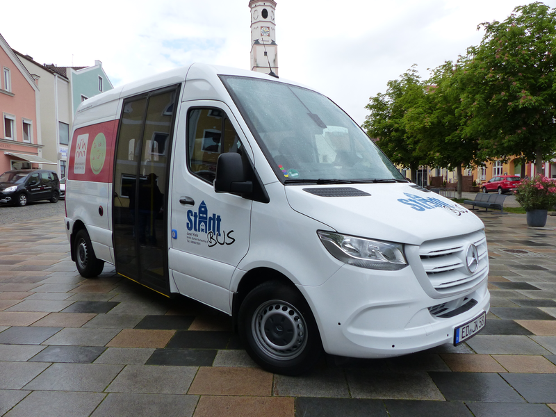 Ein weißer Kleinbus mit der Aufschrift "Stadtbus" steht auf einem gepflasterten Platz vor bunten Häusern und Bäumen.