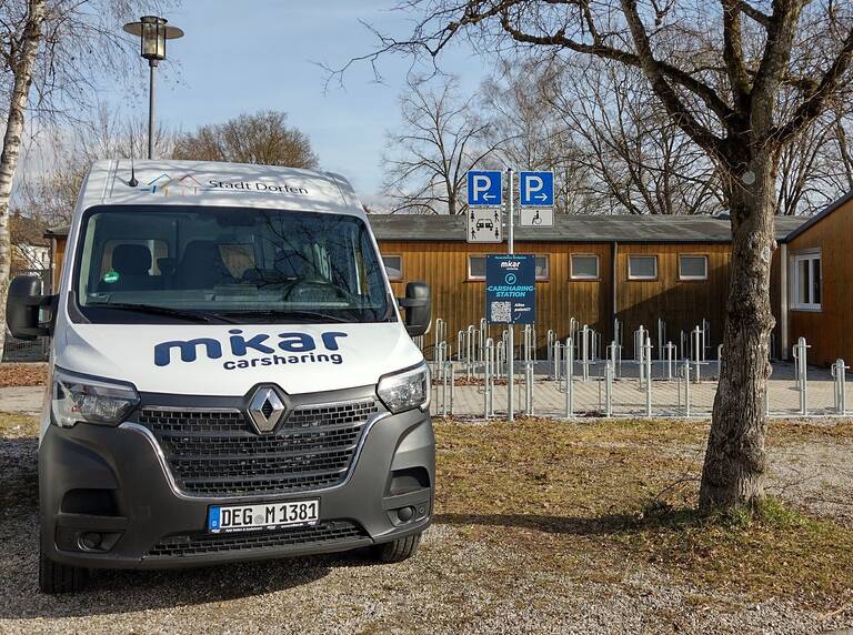 Ein Carsharing-Fahrzeug von "mikar" steht vor einer Holzhütte mit Fahrradständern und Parkschildern im Hintergrund.