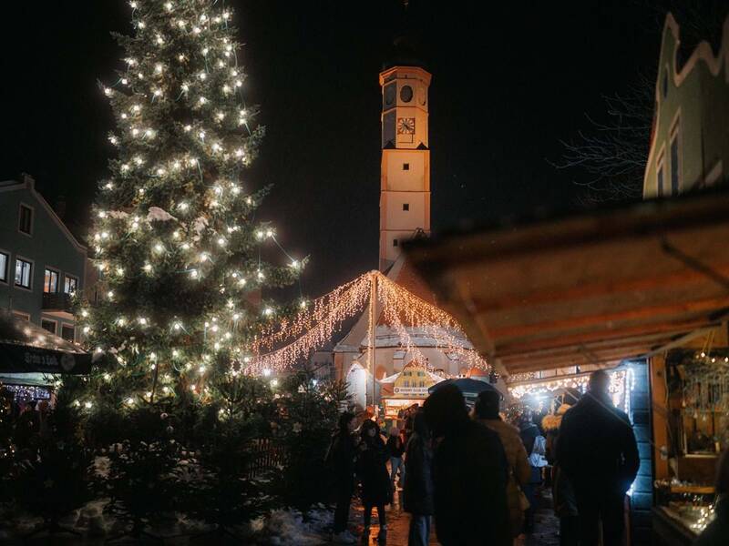 Eine weihnachtlich beleuchtete Szenerie zeigt einen großen, strahlenden Weihnachtsbaum neben einem Turm und von Lichtern geschmückte Stände auf einem Weihnachtsmarkt bei Nacht.